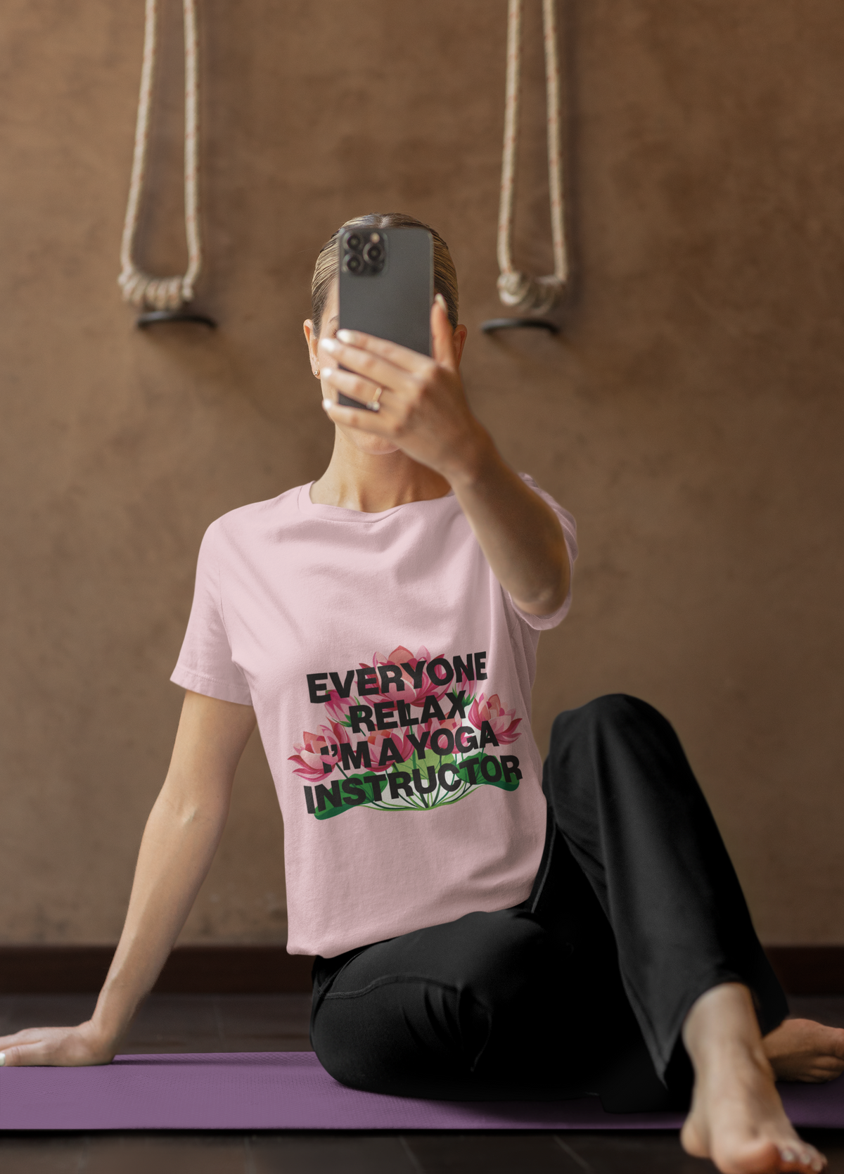 Person sitting on a yoga mat wearing a pink t-shirt with text, in a room with a brown wall and ropes.